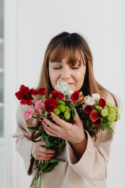 A woman with long, light brown hair and bangs, dressed in a beige sweater, is gently holding a vibrant bouquet of fresh flowers against a plain white background. The bouquet features a mix of red, pin