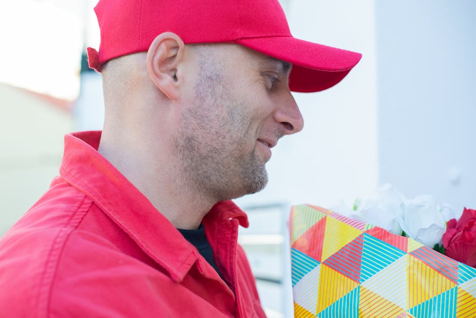 A man wearing a red baseball cap and matching red jacket is holding a colorful wrapped floral gift. The bouquet includes red and white flowers, partly visible inside a geometric-patterned paper wrappi