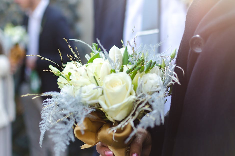 A close-up of a floral bouquet featuring cream-colored roses, white tulips, and delicate greenery arranged in a rounded, hand-held bouquet. The flowers display fresh, soft petals with a subtle sheen, 