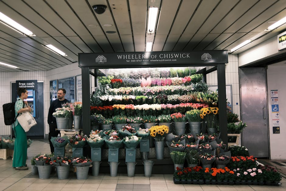 A flower stall titled 'Wheeler of Chiswick' located in a train station with a large, colorful display of fresh floral arrangements. The arrangement includes a variety of flowers such as pink roses, wh