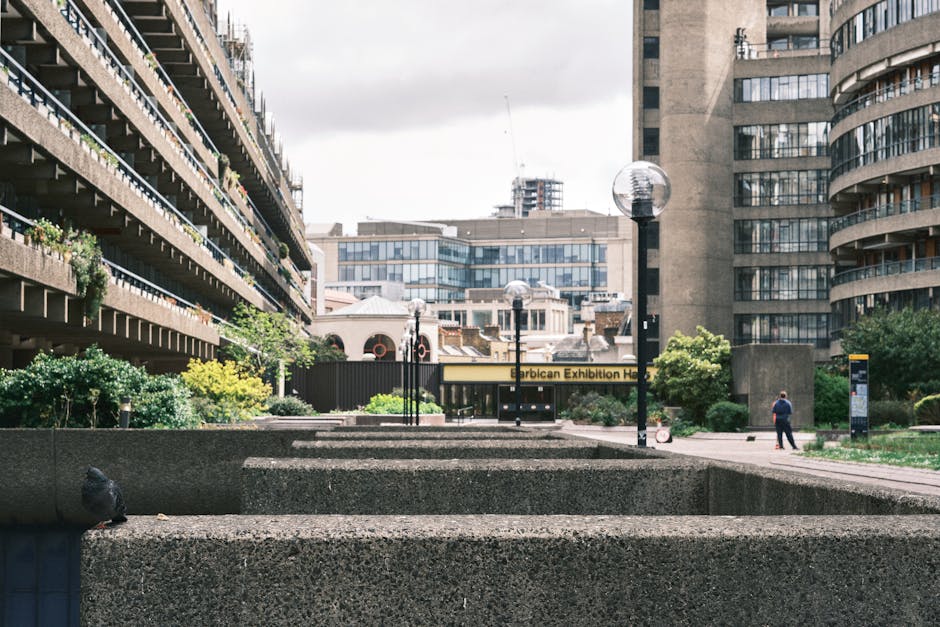 A view of the exterior courtyard of the Barbican Estate in London, featuring multi-storey concrete residential buildings on either side with rows of balconies adorned with green plants and flowers. Th
