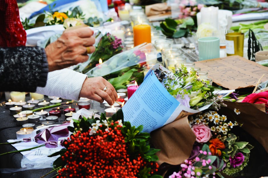 A floral tribute featuring a bouquet of mixed flowers, including pink roses, white daisies, and purple lisianthus, arranged with green foliage. The bouquet is wrapped in brown craft paper and tied wit